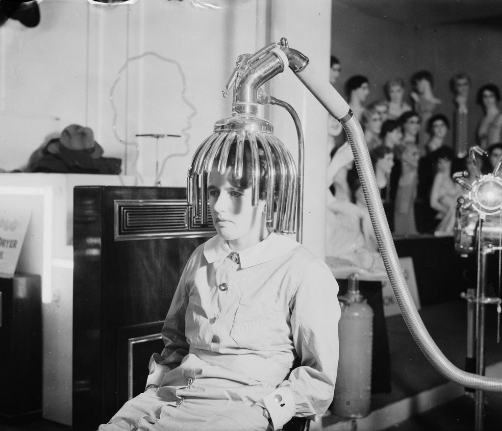 Woman sitting under a hairdryer, 9 September 1934 Black and white photograph of a woman sitting under an early hairdryer